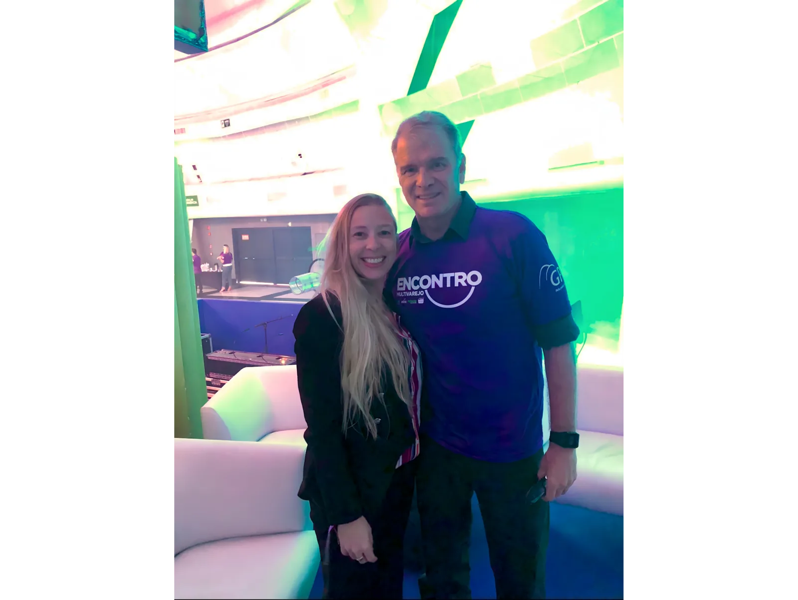 Pilar Moretzsohn e outro participante posam em um espaço de evento iluminado.