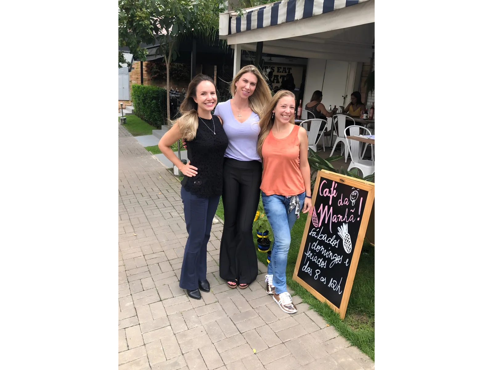Pilar Moretzsohn posa com outras duas mulheres na área externa de um café.